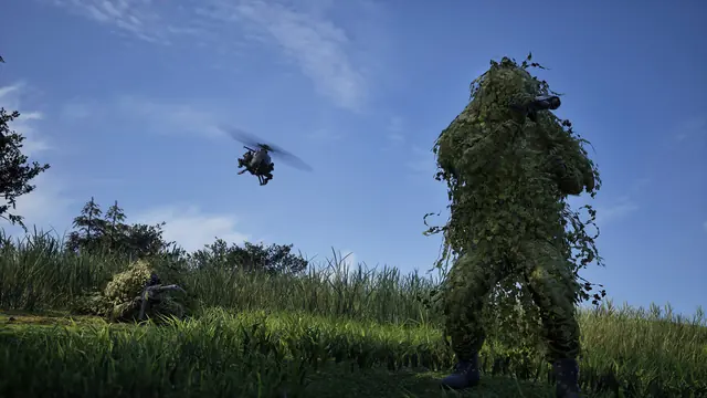 A sniper in a full ghillie suit stands in tall grass as a helicopter passes